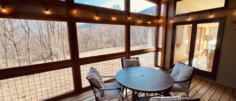 Eine Holzterrasse mit rundem Tisch und Stühlen, große Fenster mit Lichterketten und Blick auf ein bewaldetes Gebiet.