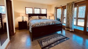 A bedroom with a large bed, two bedside tables, a nightstand, a window with curtains, and a view of trees.