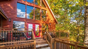 A wooden cabin with a red door, a deck with outdoor seating, and a staircase leading to the entrance. - Where Mountain views and quiet moments come naturally, it’s s’more than enough (Sevierville)