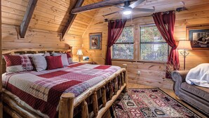 A cozy log cabin bedroom with a wooden bed, a nightstand, a lamp, a window with curtains, and a view of the outdoors. - Where Mountain views and quiet moments come naturally, it’s s’more than enough (Sevierville)