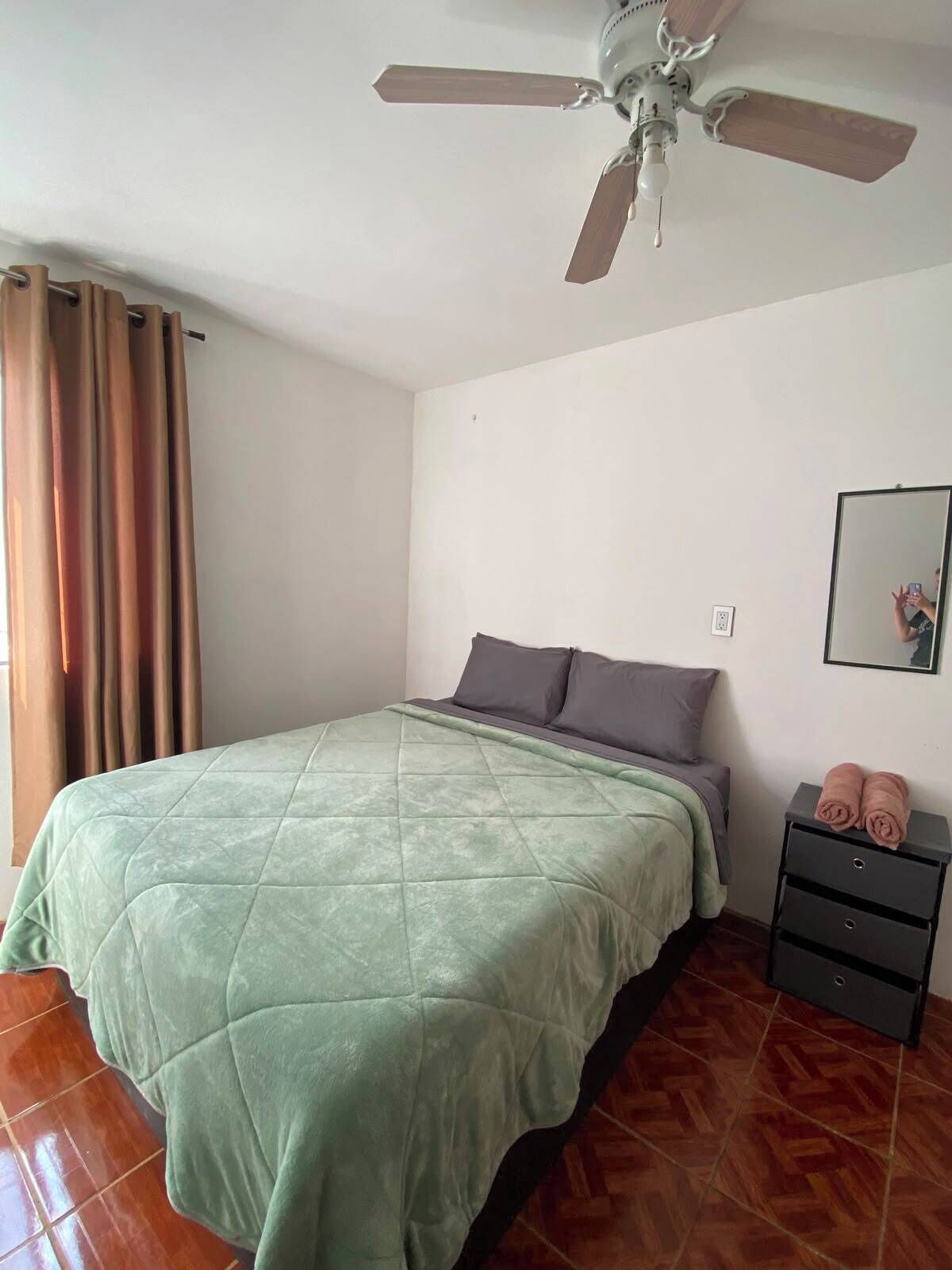 Una camera da letto con un letto, una ventola a soffitto, un comodino con uno specchio e una finestra con tende.