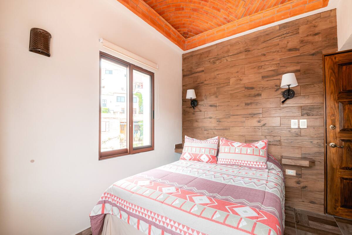 Un dormitorio con cabecera de madera, una cama grande con ropa de cama estampada, dos lámparas fijadas a la pared y un ventanal con vistas a edificios.