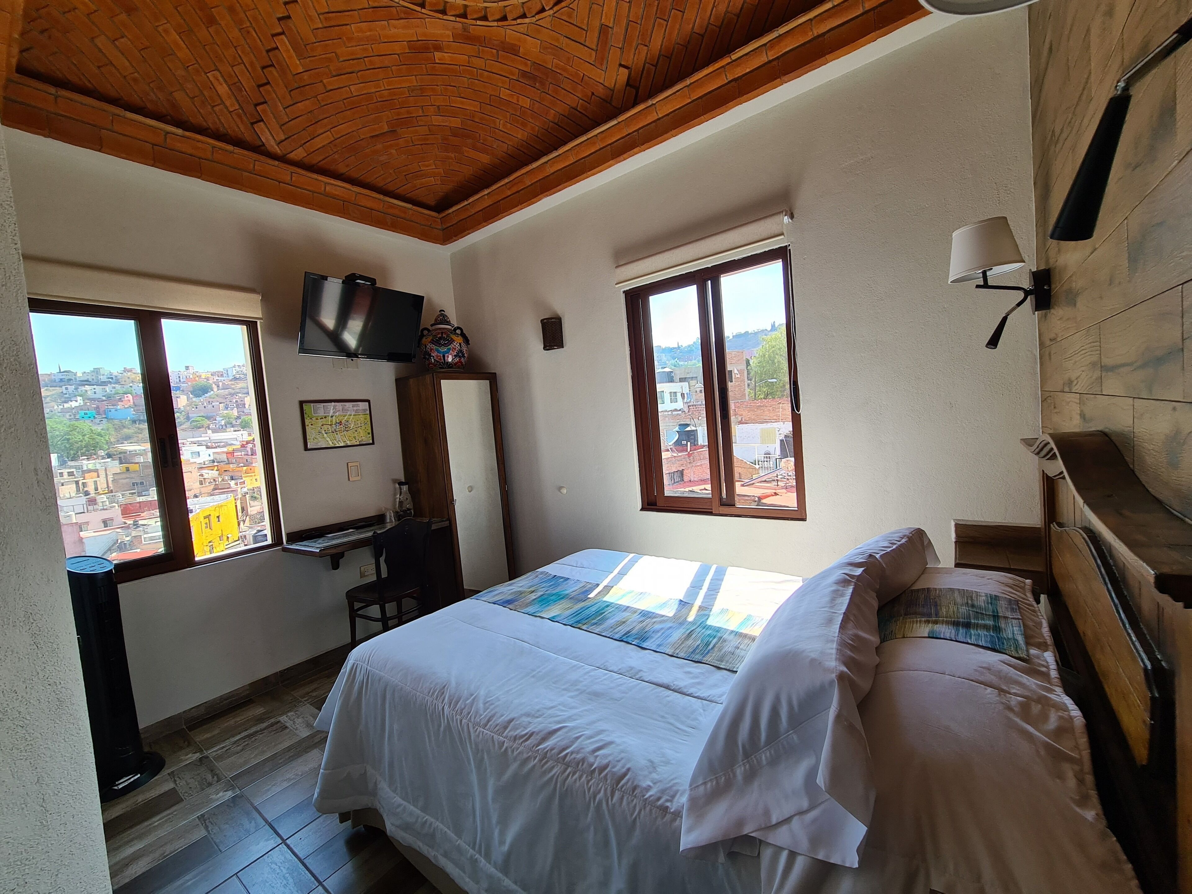 Un dormitorio con cama, escritorio, silla, televisor y ventana con vista a la ciudad.