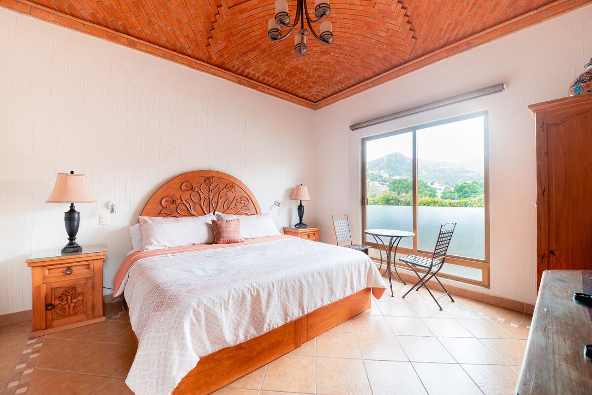 Un dormitorio con una cama grande, dos mesitas de noche, un cabecero de madera, vistas al exterior a través de un ventanal amplio y una pequeña mesa con sillas.