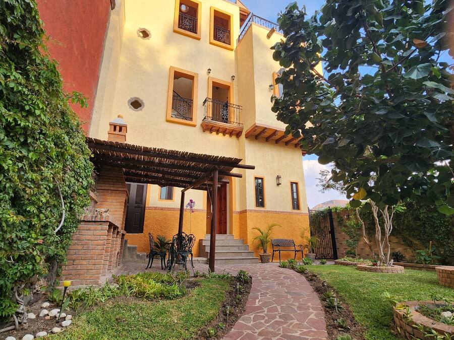 Um edifício de dois andares com fachada amarela, um caminho de pedra que leva à entrada e um pequeno pátio com mobiliário de exterior.