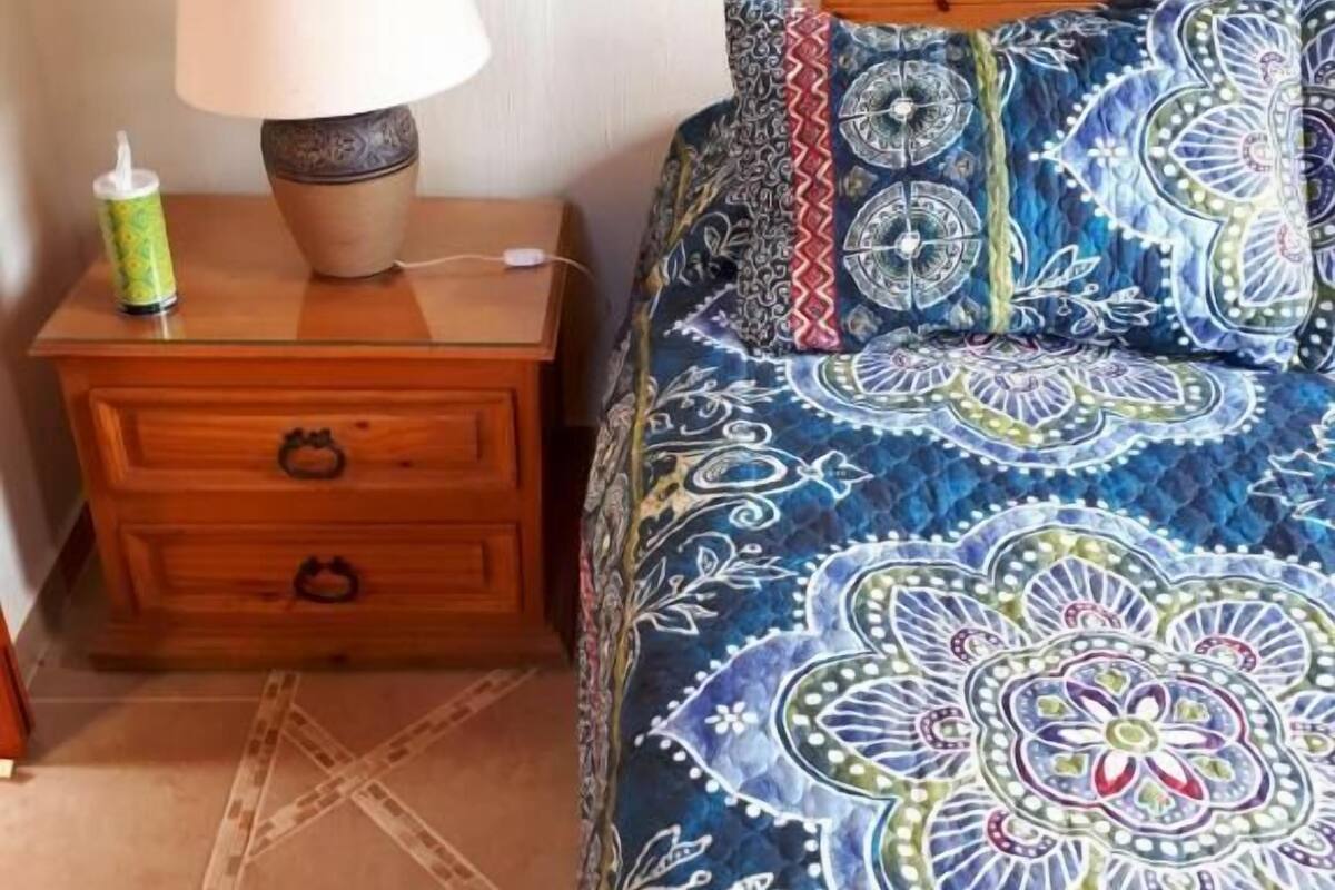 Un dormitorio con una mesita de noche de madera, una lámpara, una colcha estampada y un cojín decorativo.