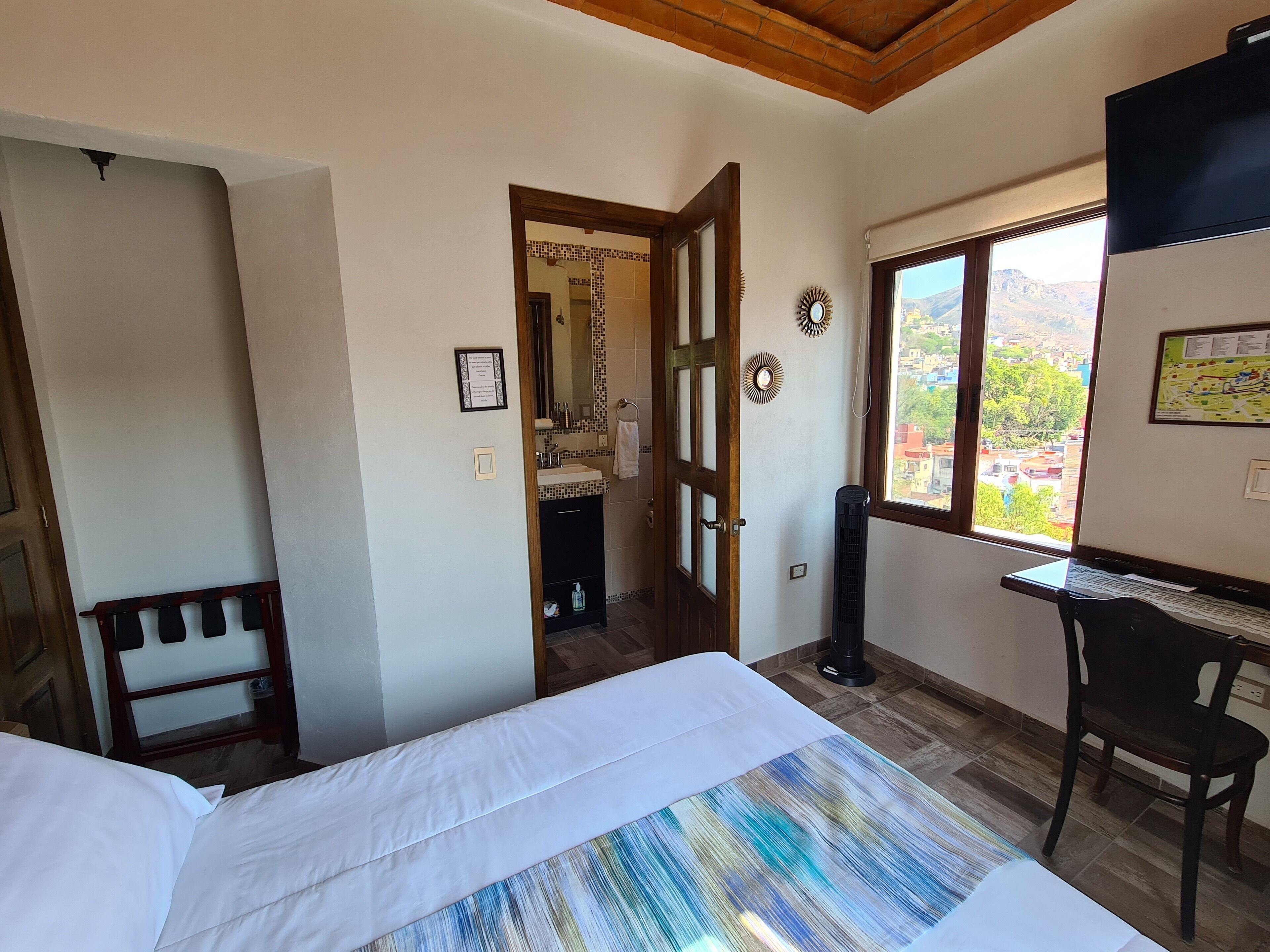 A bedroom with a bed, a desk, a chair, a window with a view, and a door leading to another room.