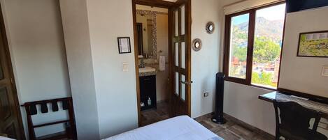 A bedroom with a bed, a desk, a chair, a window with a view, and a door leading to another room.