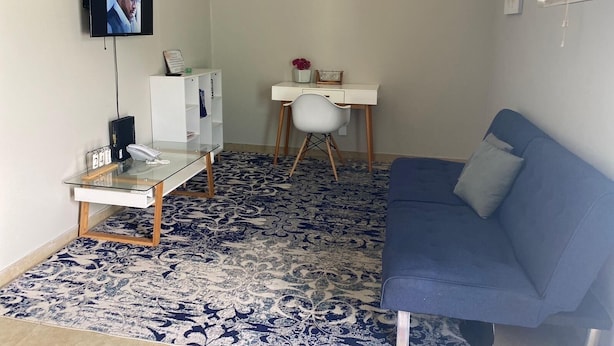 Una habitación con un sofá azul, un escritorio blanco, una mesa de vidrio y una alfombra estampada.