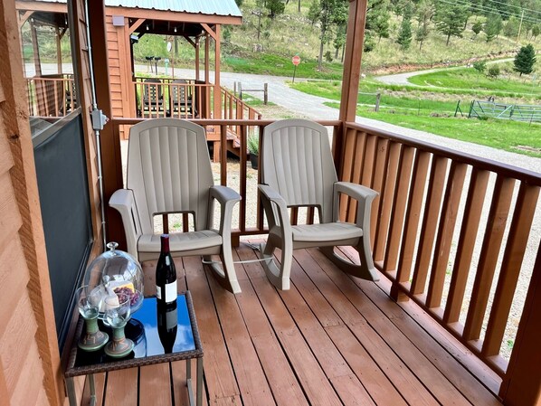 A wooden deck with two chairs and a table, a stop sign, and a view of a green landscape. - Tiny Cottages In Cloudcroft / Mayhill NM (Mayhill)