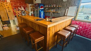 Una zona de bar con mostrador de madera, taburetes, refrigerador de Pepsi, estantes con varias botellas y vistas al exterior a través de una ventana.