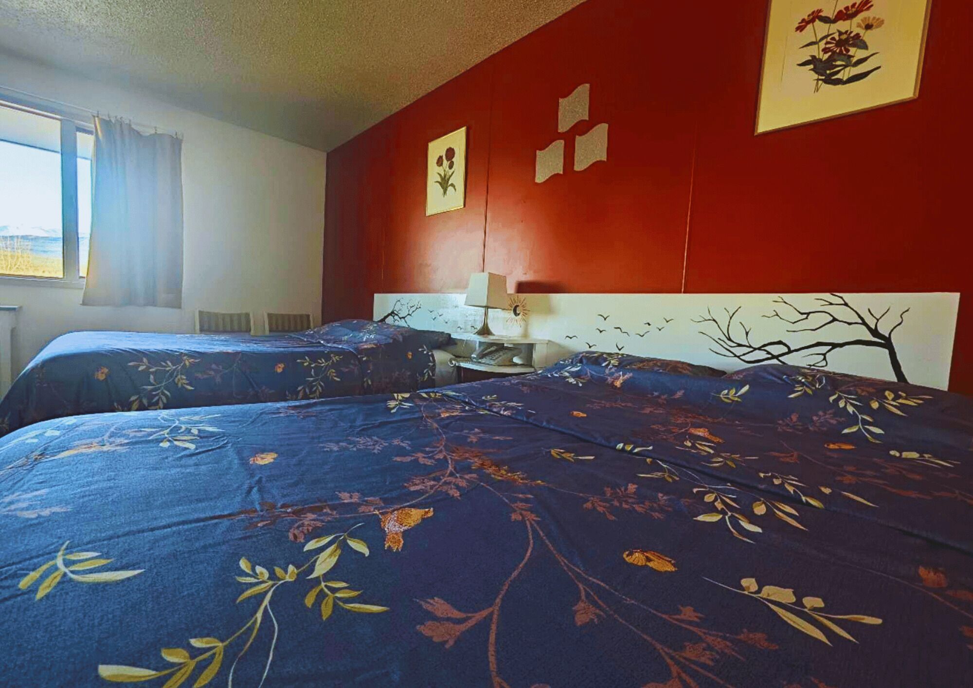 Ein Zimmer mit zwei Betten, jeweils mit floralem Bettbezug, einer roten Wand und einem Fenster mit Vorhängen.