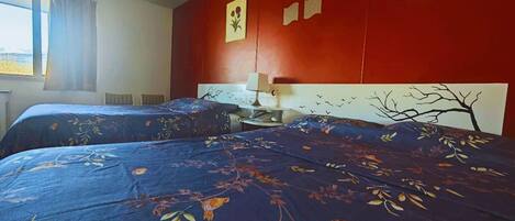 Ein Zimmer mit zwei Betten, jeweils mit floralem Bettbezug, einer roten Wand und einem Fenster mit Vorhängen.