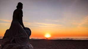 Eine Statue auf einer gewölbten Plattform mit Blick auf einen Strand bei Sonnenuntergang.