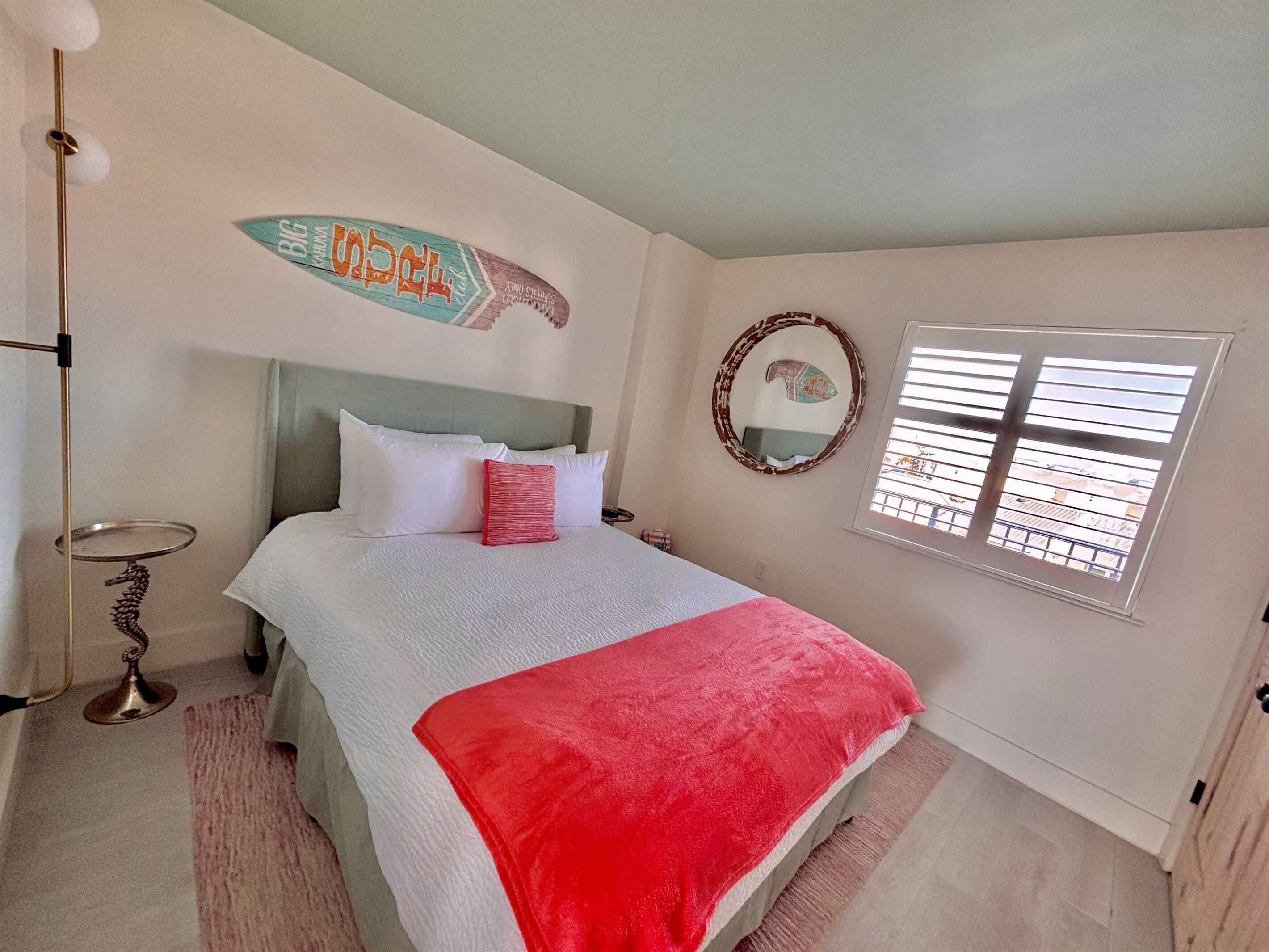 A bedroom with a bed, a bedside table, a mirror, and a window with blinds.