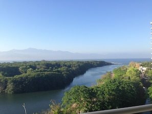 Une rivière bordée d’une végétation luxuriante, avec des montagnes en arrière-plan et un ciel dégagé.