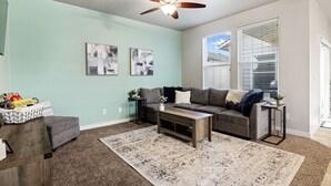 A living room with a gray sofa, a coffee table, and a ceiling fan. There are two abstract paintings on the wall and a window with blinds. - ❃Close to the POOL! 3BD/2BA! Close to BSU/Nampa/Kuna! Your Boise Vacation Spot!❃ (Meridian)