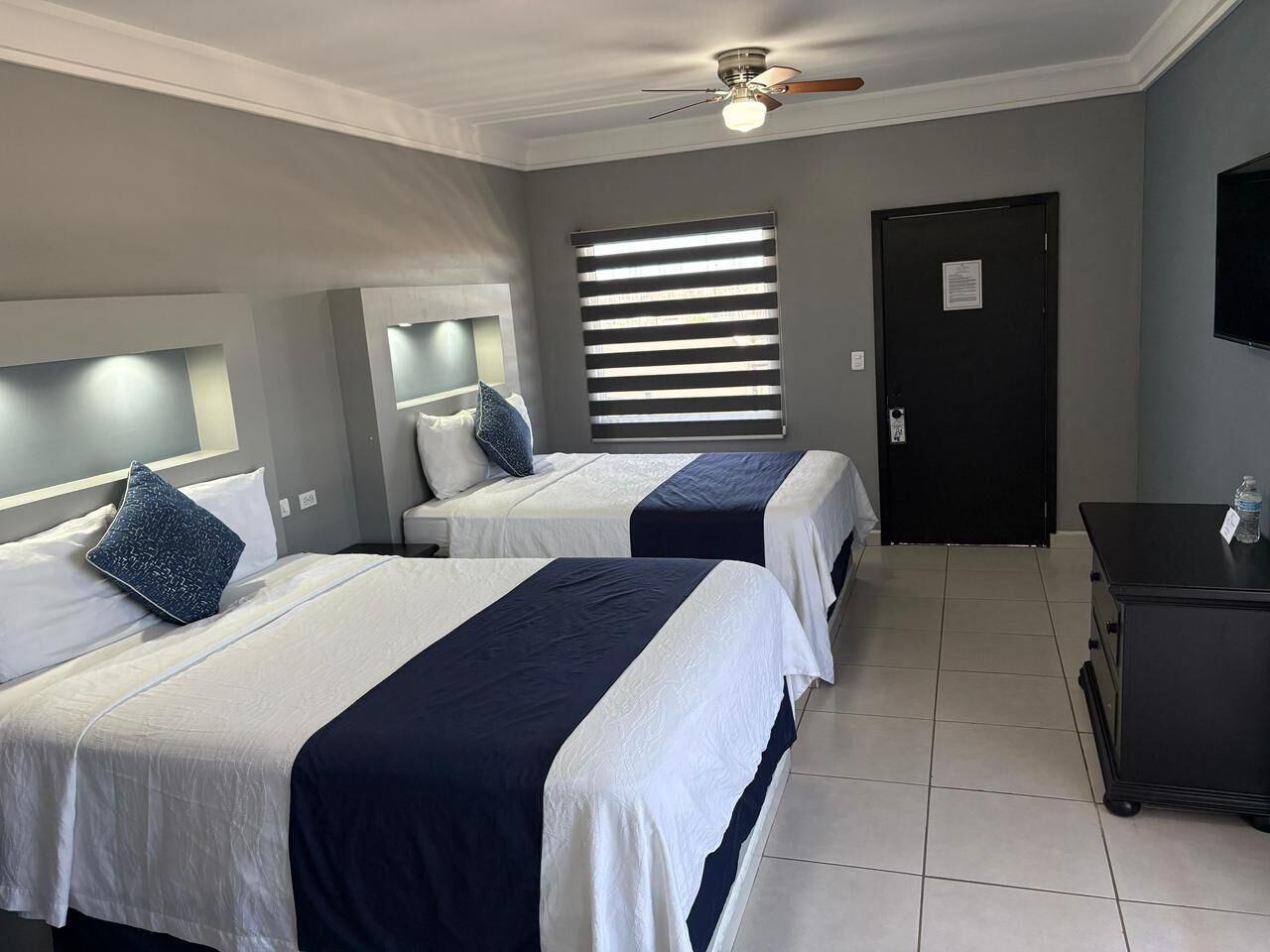 A hotel room with two beds, a ceiling fan, a window with blinds, a door, and a small table with a bottle.