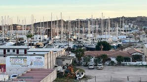 Un puerto deportivo con numerosas embarcaciones amarradas, un cartel publicando eventos locales y algunos coches estacionados cerca.