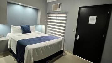 Ein Hotelzimmer mit Bett, Deckenventilator, Fenster mit Jalousien, einer Tür und einem kleinen Tisch mit Lampe.