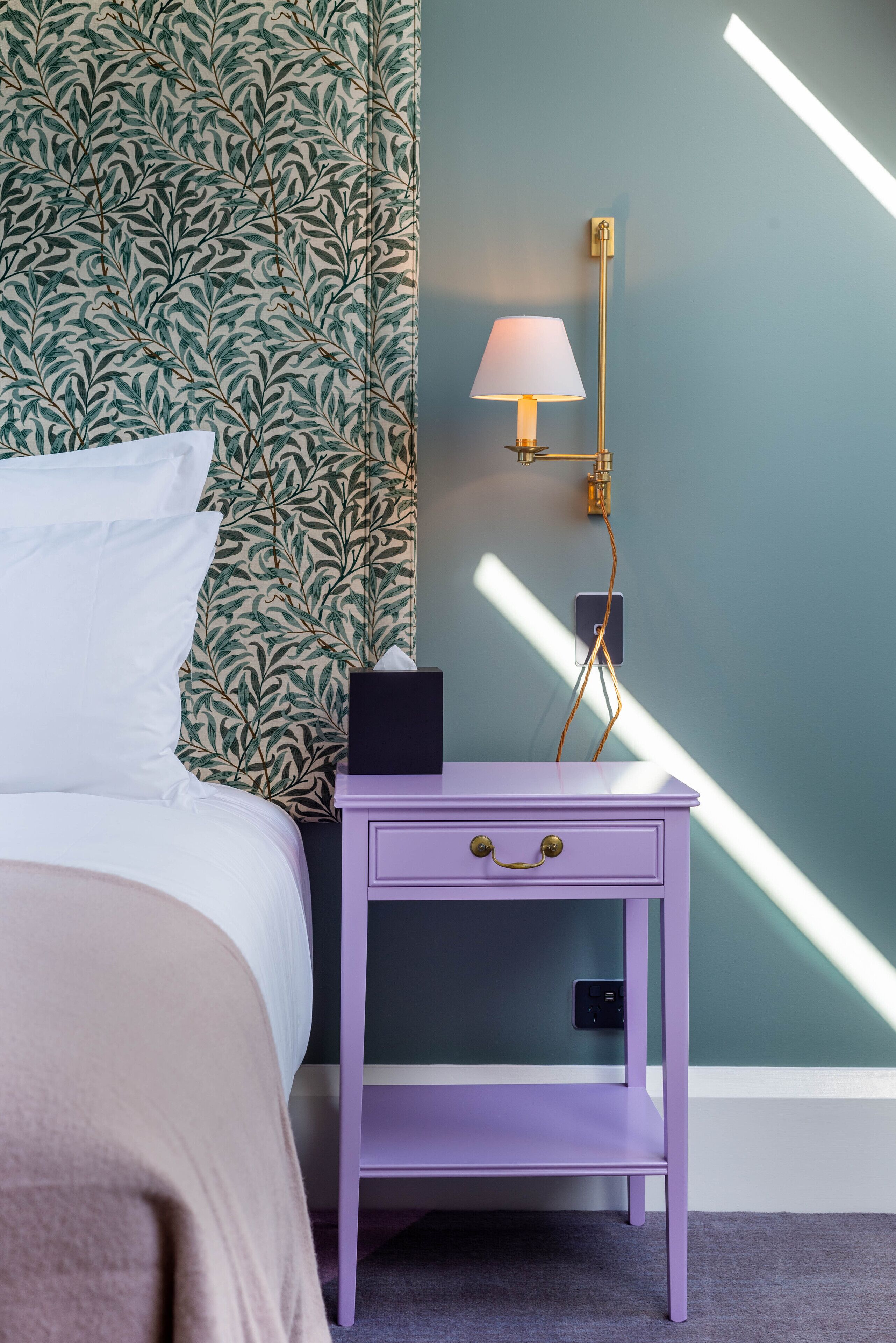 Un lit bien fait, avec une literie blanche, une table de chevet violette surmontée d’une lampe, et un mur orné d’un motif végétal.