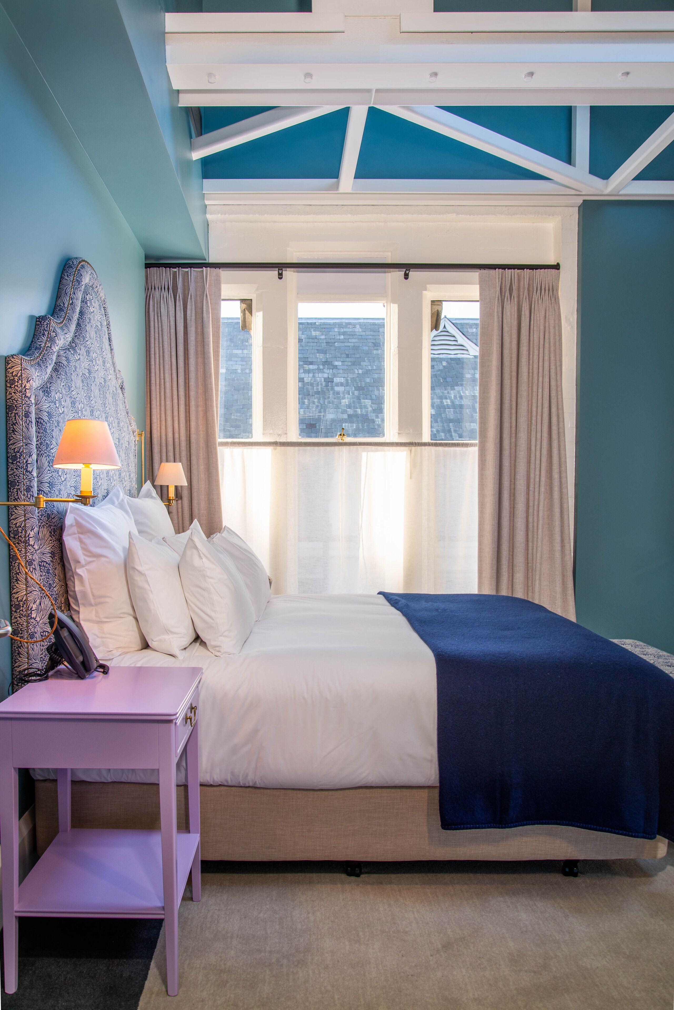 A bedroom with a bed, bedside lamps, a nightstand, and a view of the city through large windows.