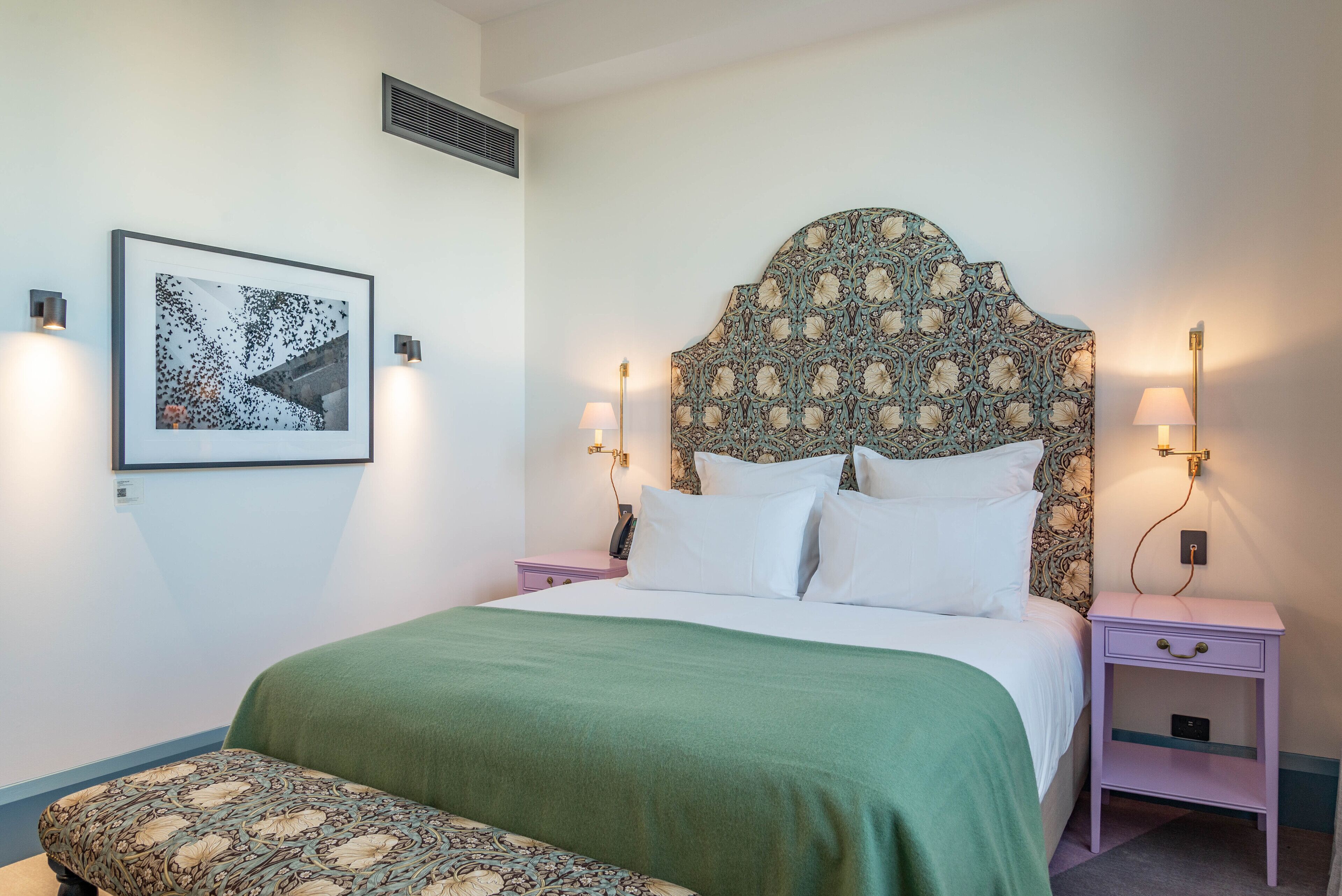 A neatly arranged bedroom with a large bed, a bedside table, a nightstand, a bench, and a wall-mounted picture.