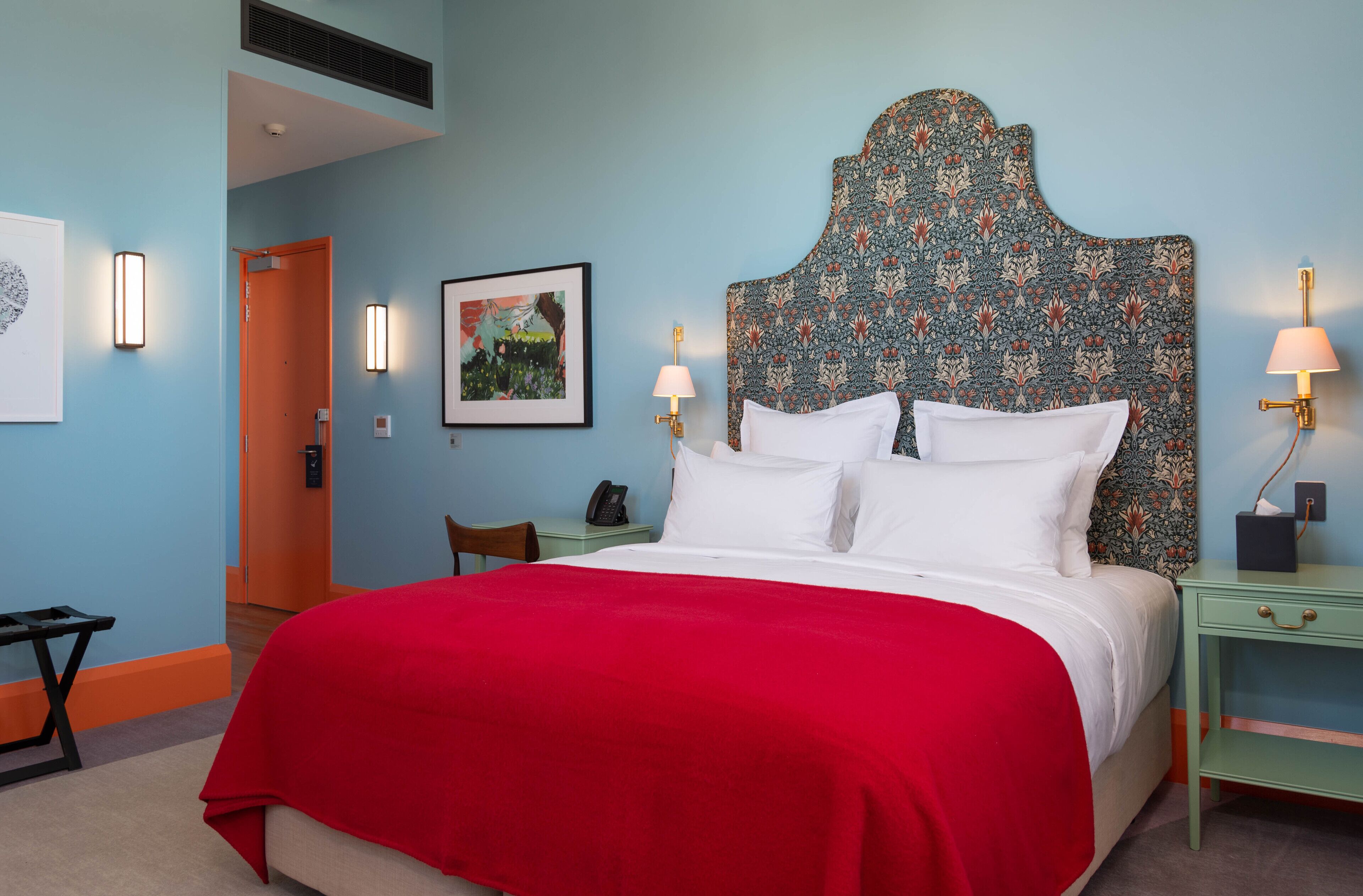A hotel room with a large bed, a red bedspread, two bedside lamps, a nightstand, a framed picture on the wall, and a door leading to another room.