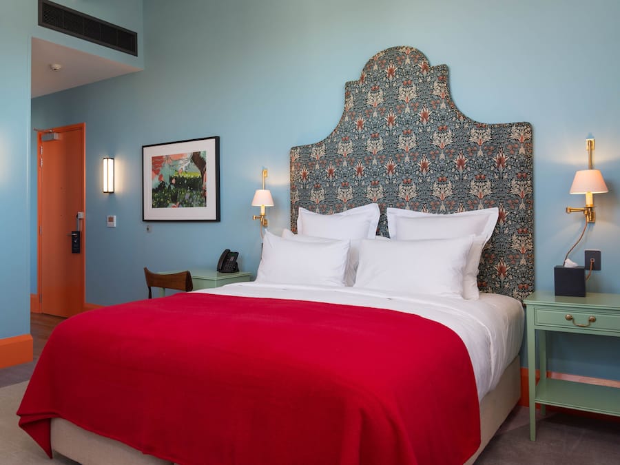 A hotel room with a large bed, a red bedspread, two bedside lamps, a nightstand, a framed picture on the wall, and a door leading to another room.