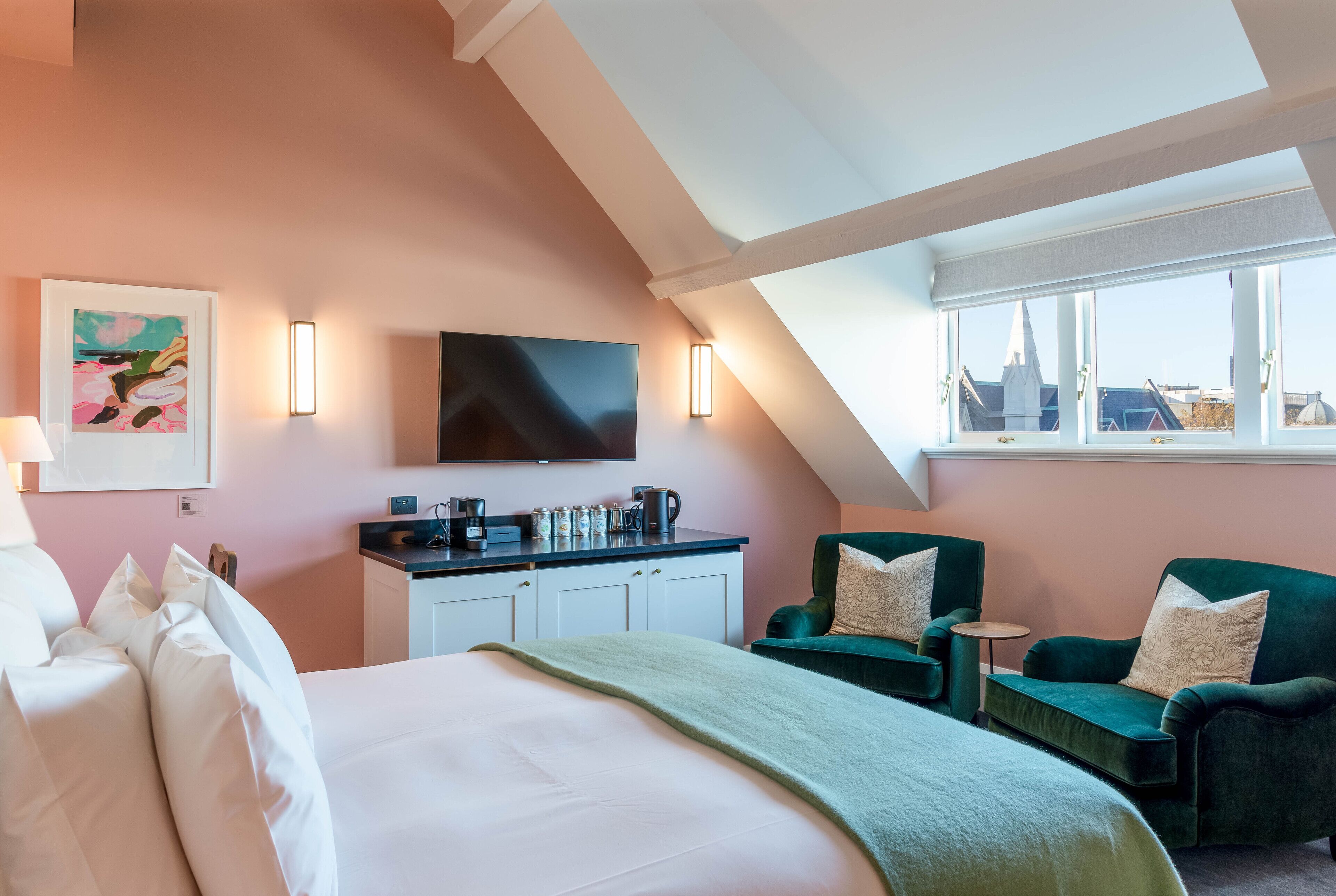 A hotel room with a bed, two green armchairs, a small table, a flat-screen TV, and a city view through large windows.