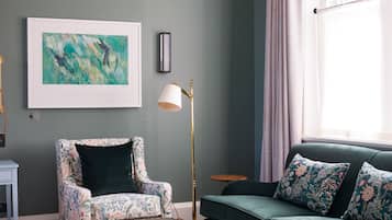 A living room with a green sofa, a floral armchair, a glass coffee table, and a framed abstract painting on the wall.