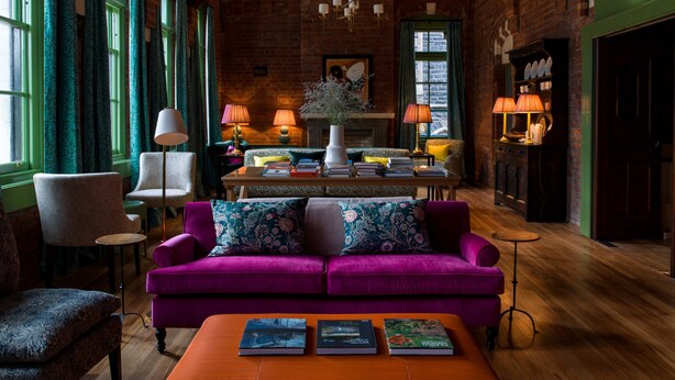 Une pièce chaleureuse avec un canapé violet, un plancher en bois et un mur en briques. On y trouve une table avec des livres, une lampe et une cheminée.