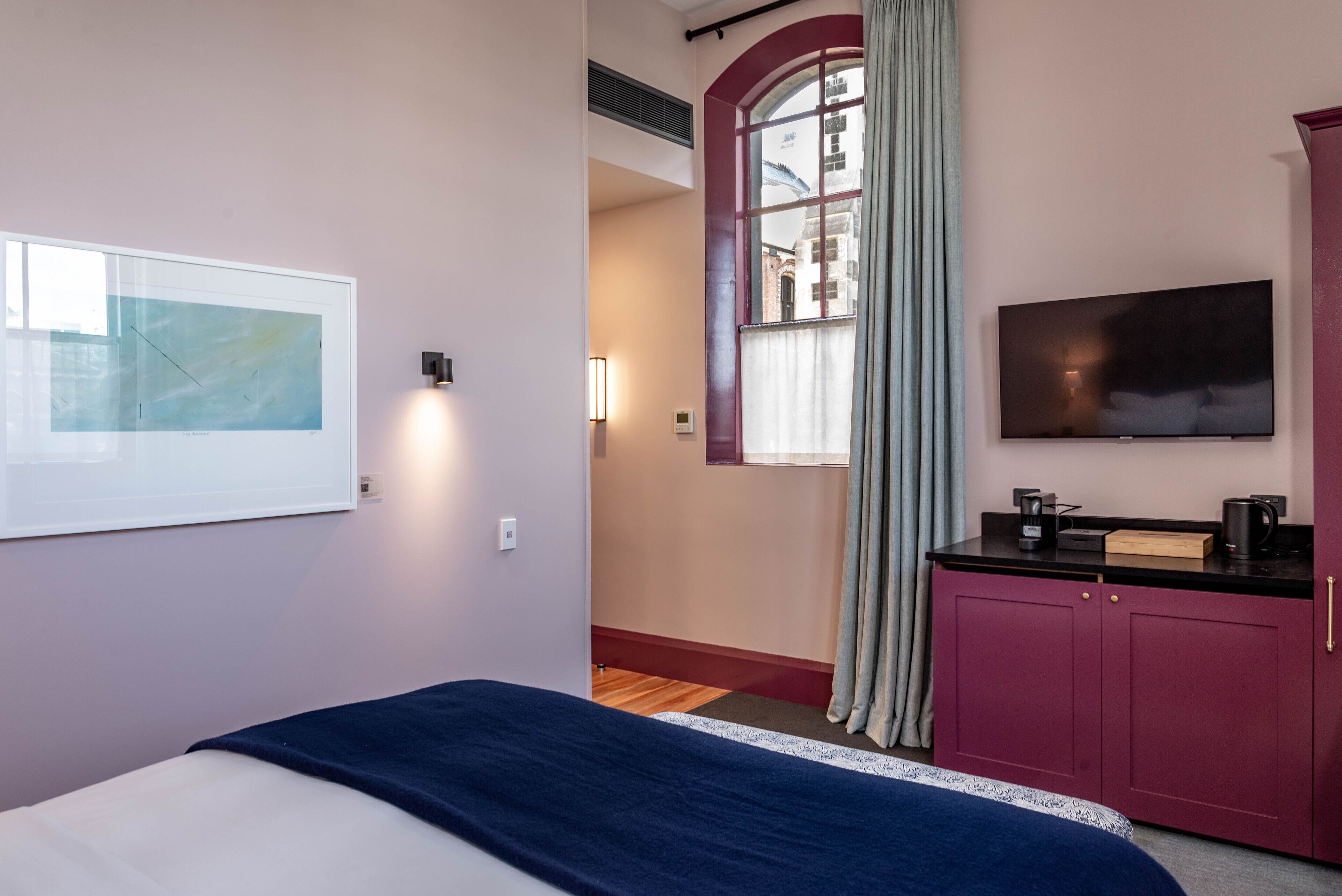 Une chambre d’hôtel avec un lit, une télévision, un tableau encadré, une fenêtre avec des rideaux et une armoire rouge.