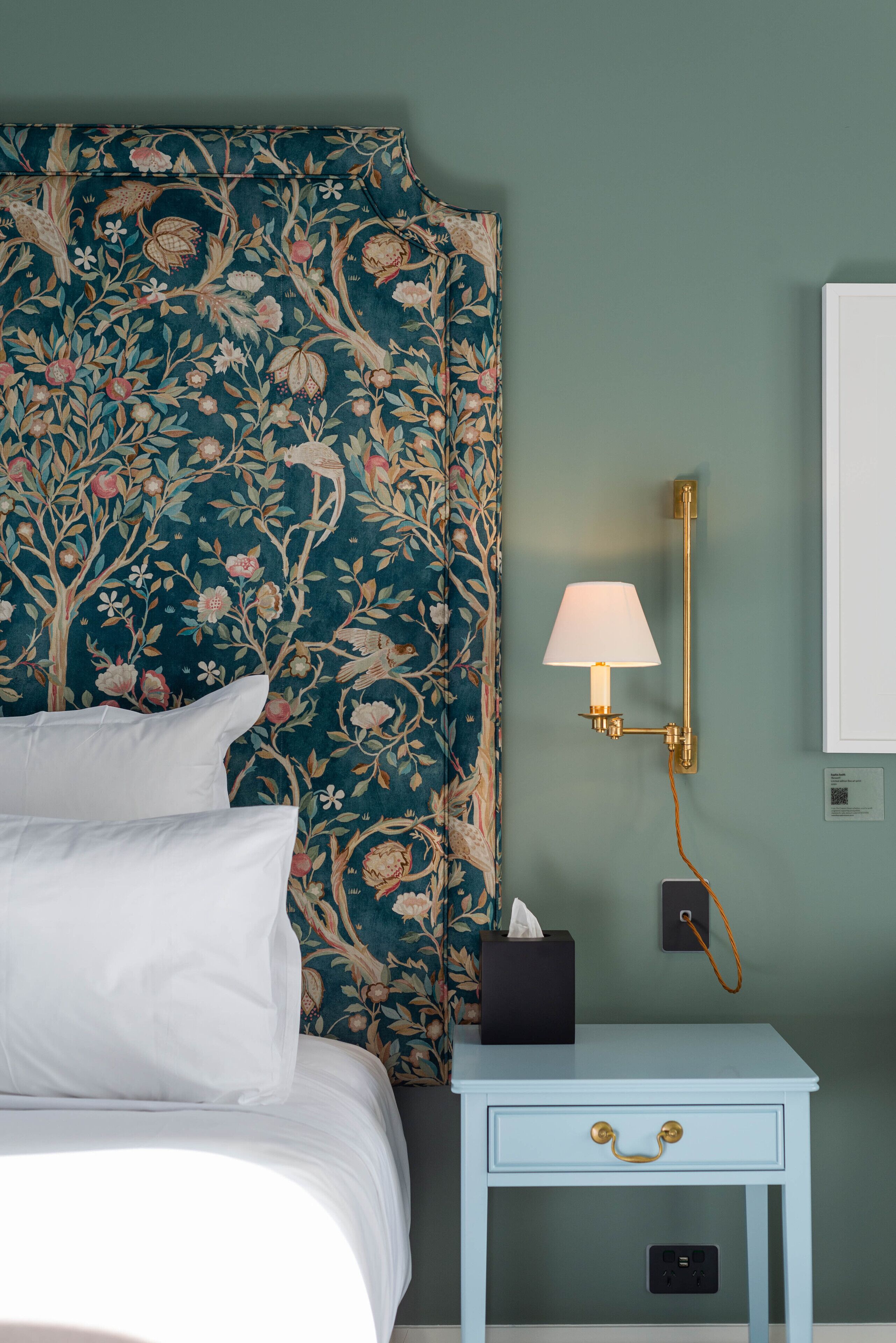 A neatly made bed with white linens, a floral-patterned headboard, a bedside table with a lamp, and a small black box.