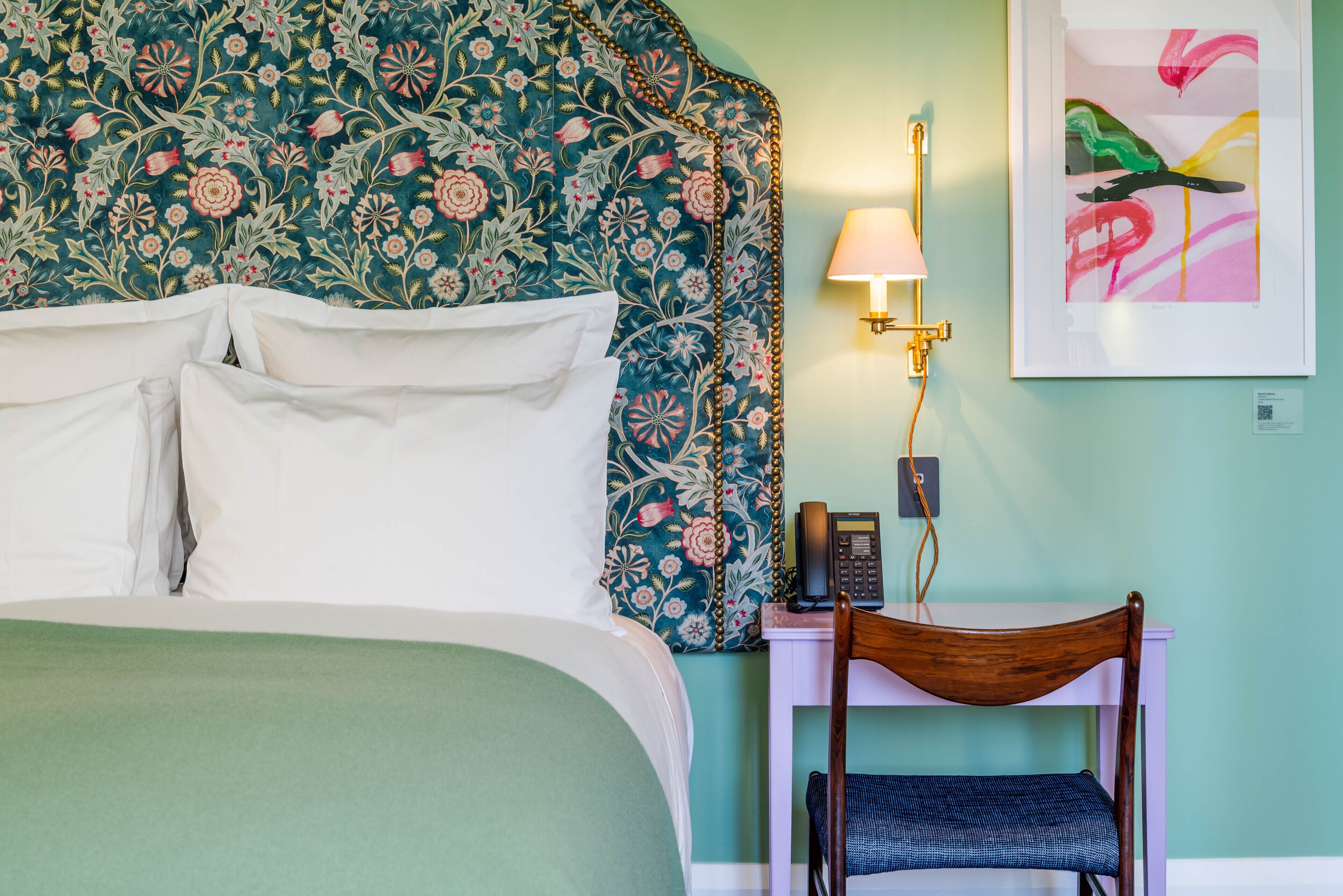 Una cama bien hecha con sábanas blancas, un cabecero floral, una mesita de noche con una lámpara, una silla y una obra de arte enmarcada en la pared.