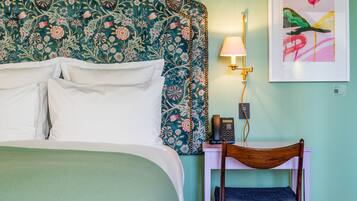 Una cama bien hecha con sábanas blancas, un cabecero floral, una mesita de noche con una lámpara, una silla y una obra de arte enmarcada en la pared.