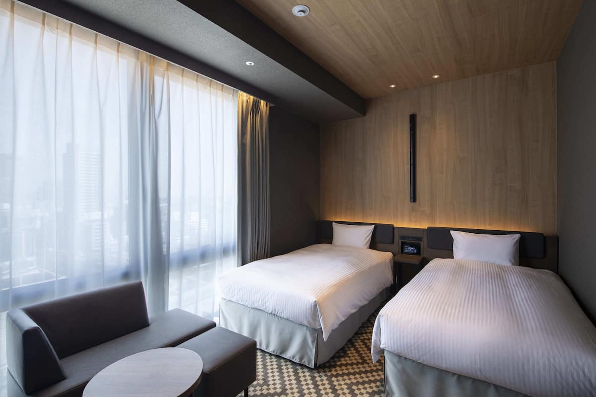 Een hotelkamer met twee bedden, een sofa, een kleine tafel en uitzicht op de stad door grote ramen.