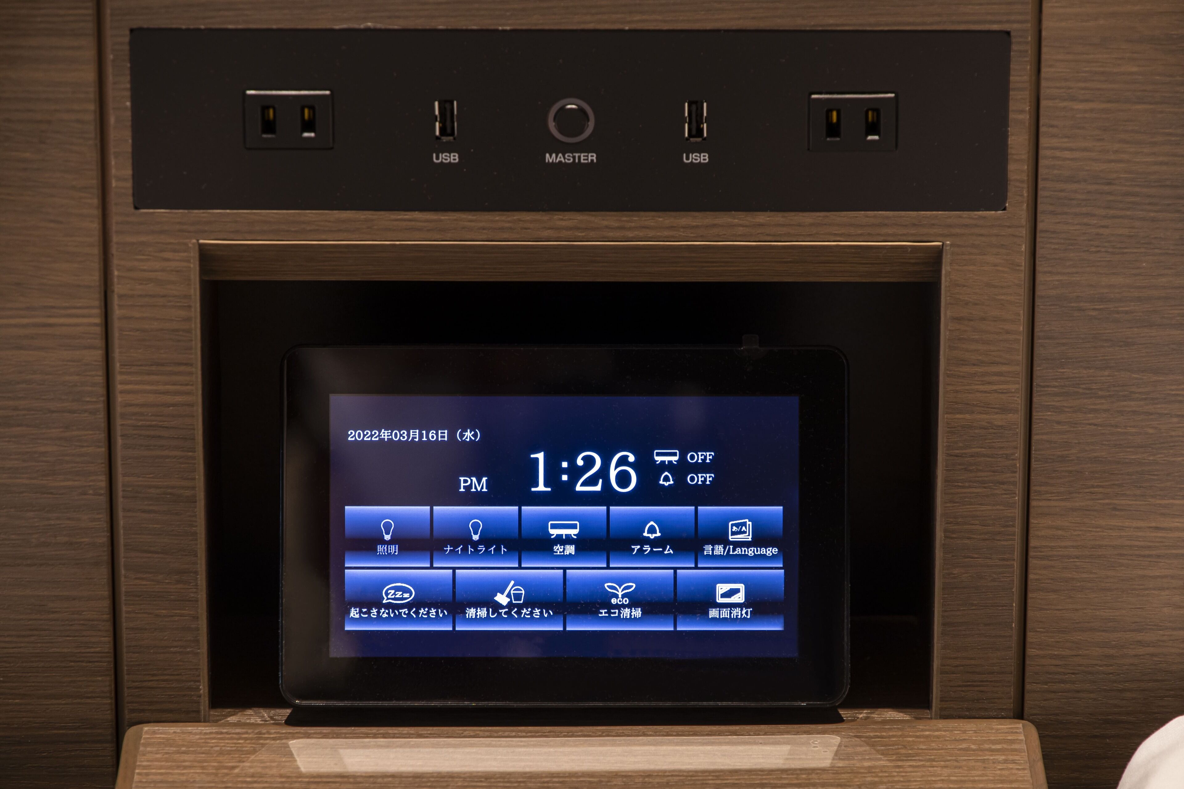 Pantalla digital en un panel de madera que muestra fecha, hora y varios iconos para controlar las funciones de la habitación.