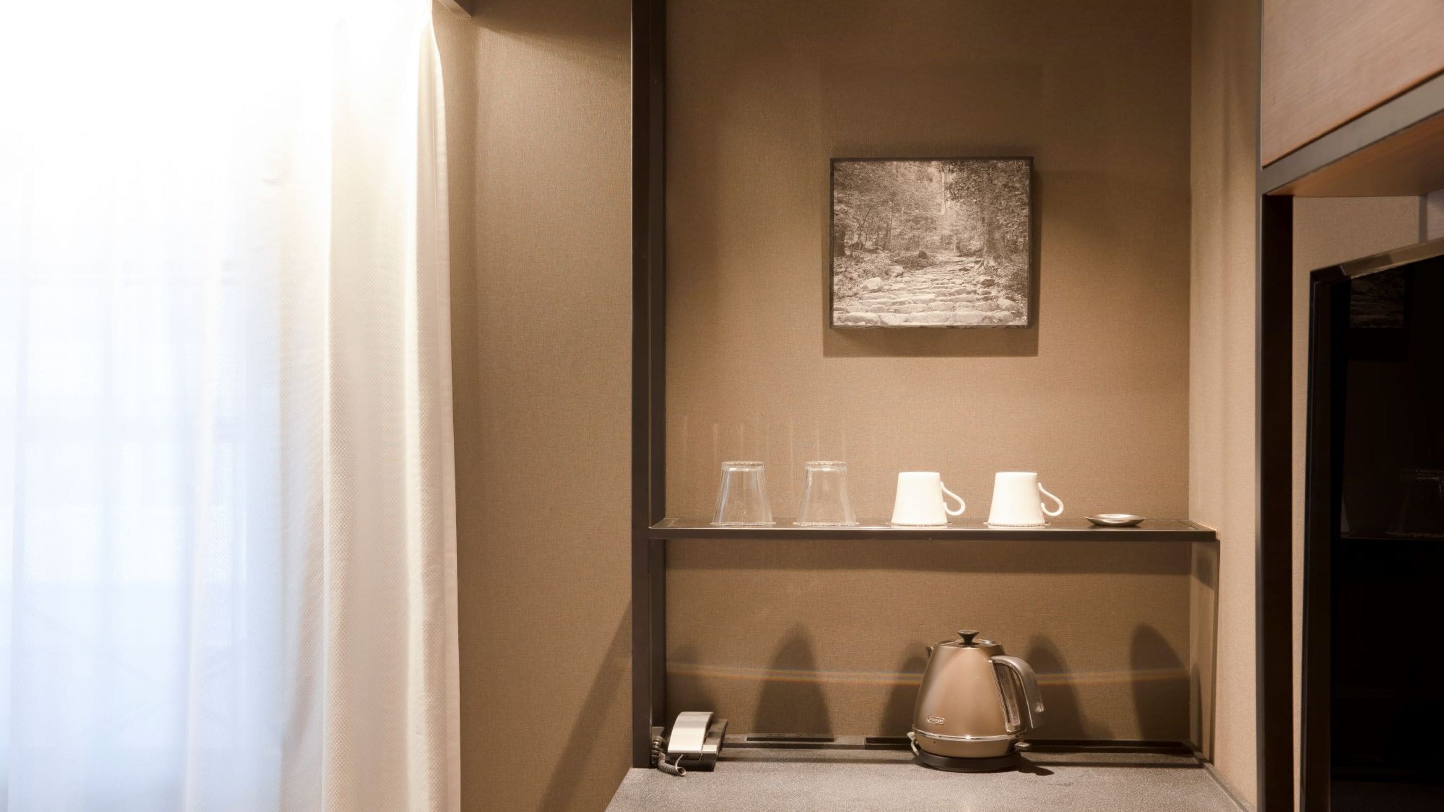 A hotel room with a beige wall, a framed picture, a shelf with cups, a kettle, and a telephone.