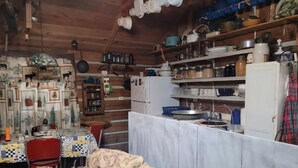 A rustic cabin interior with a wooden ceiling, a dining area with a checkered tablecloth, a sofa, and a kitchen with shelves and a refrigerator.
