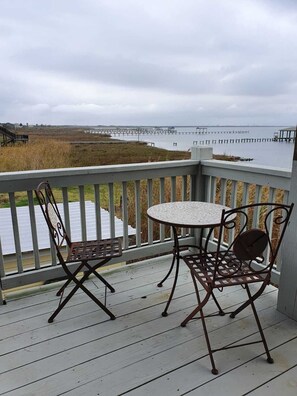 A wooden deck with a table and chairs, overlooking a body of water and a distant pier.