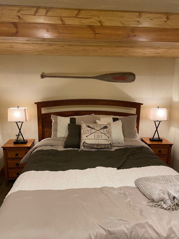 A bedroom with a wooden ceiling, a bed with a quilted bedspread, two bedside lamps, and a pillow with a canoe and text.