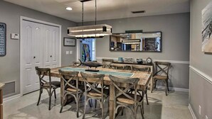 A dining area with a wooden table and chairs, a hanging light fixture, and a kitchen area in the background.