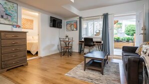 A compact living space with a wooden floor, a sofa, a desk with a chair, a TV, and a dining area with a table and chairs. - Private Suite!! Convenient location for Commuters, Essential Workers and Trades! (New Westminster)