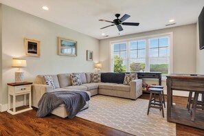 Una sala de estar con un sofá modular, un ventilador de techo, una ventana con persianas y una pequeña mesa con taburetes.