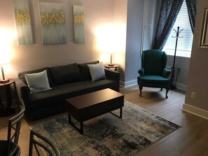 Een zithoek met een donkerblauwe bank, een houten bijzettafeltje en een blauwgroene fauteuil. Aan de muur hangen abstracte schilderijen en er staat een vloerlamp.