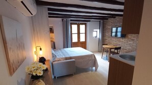Un dormitorio acogedor con una cama, una mesita de noche y un jarrón de flores. Se observa una pequeña kitchenette con un lavabo y una ventana al fondo.
