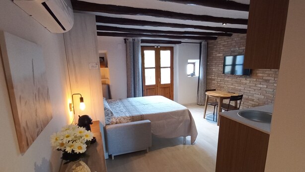 A cozy bedroom with a bed, bedside table, and a vase of flowers. A small kitchenette with a sink and a window is visible in the background.