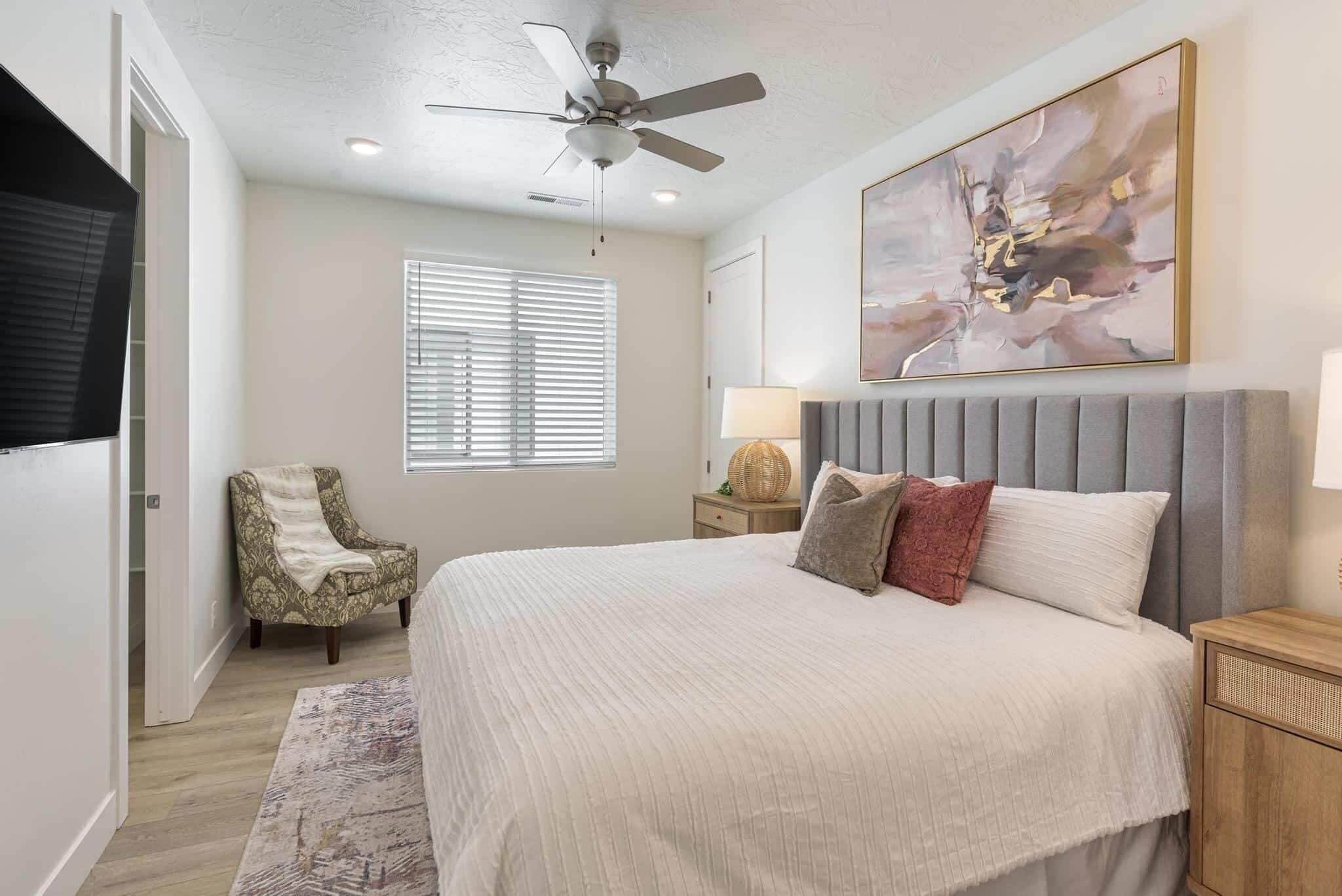 A bedroom with a bed, a chair, a ceiling fan, a window with blinds, a lamp, and a painting on the wall.