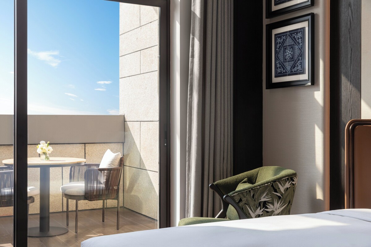 A modern hotel room with a large window, a balcony with a table and chairs, and a green upholstered chair.